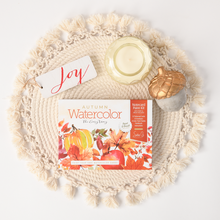 A flat lay of autumn-themed items on a round woven mat: Schiffer Publishing, Ltd.'s Watercolor the Easy Way Paint-Your-Own Watercolor Cards Autumn kit, a white candle, a ceramic acorn, and a white tag with "Joy" in red script.