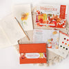 A flat lay of Schiffer Publishing, Ltd.'s Watercolor the Easy Way Paint-Your-Own Watercolor Cards Autumn kit, featuring a paint palette, instruction booklet, brush, floral notecards, and orange envelope on a round woven mat.