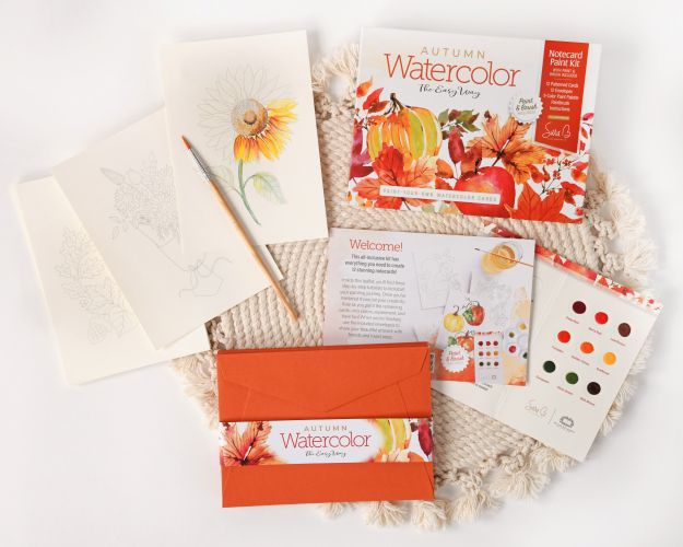 A flat lay of Schiffer Publishing, Ltd.'s Watercolor the Easy Way Paint-Your-Own Watercolor Cards Autumn kit, featuring a paint palette, instruction booklet, brush, floral notecards, and orange envelope on a round woven mat.