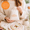A woman in a cream sweater holds the Sunny Stitches Embroidery Kit Brilliant Butterfly by Schiffer Publishing, Ltd. On the table are a butterfly hoop, flowers, a cup, stitching supplies, and orange text listing kit contents.