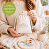 A woman holds a drawstring canvas bag labeled “Sunny Stitches Embroidery Kit Sunny Days Ahead” from Schiffer Publishing, Ltd. at a table with an embroidery hoop, needle, floss, and a finished “Sunny Days Ahead” floral embroidery.