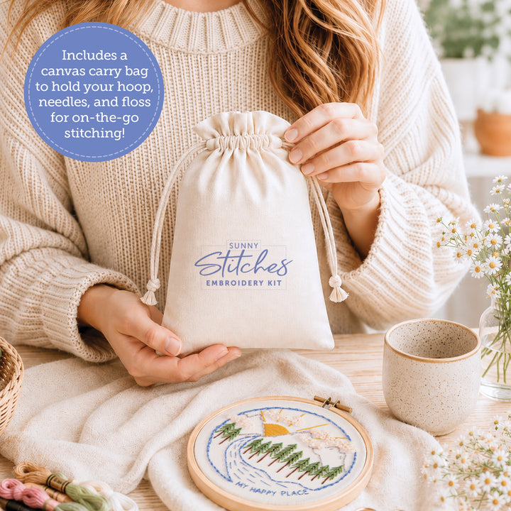 A person in a cream sweater holds a drawstring bag labeled "Sunny Stitches Embroidery Kit Sunshine Pines" by Schiffer Publishing, Ltd. On the table: a cup, flowers, and an embroidery hoop with nature design. A circle lists bag, hoop, needles, and floss inside.