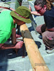 Two people work outdoors on a sustainable project inspired by "Natural Building" from Schiffer Publishing, Ltd., measuring a wooden log with pencil and string. One wears green; the other has sunglasses and dreadlocks. Others and tools are visible in the background.