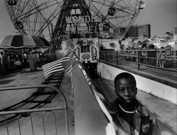 A young boy stands by a barrier at Coney Island, with a Ferris wheel and carnival booths in the background, captured in striking black and white. From Schiffer Publishing, Ltd.