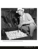 A man in a hat and sweater, pipe in hand, kneels by an open crate of golf balls, smiling with Golden Age Hollywood charm. Golf clubs are in the background. Product: Fade to Black by Schiffer Publishing, Ltd.