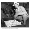 A man in a hat and sweater, pipe in hand, kneels by an open crate of golf balls, smiling with Golden Age Hollywood charm. Golf clubs are in the background. Product: Fade to Black by Schiffer Publishing, Ltd.