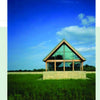 A modern glass building with a triangular roof showcases Schiffer Publishing, Ltd.'s Award-winning Green Roof Designs, standing in a grassy field beneath a blue sky and clouds, with trees lining the horizon.