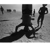 A person balances on their head in the sand at Coney Island, casting a shadow, while children walk nearby under a bright sky—an image from Schiffer Publishing, Ltd.'s "Coney Island," evoking classic black and white photography.