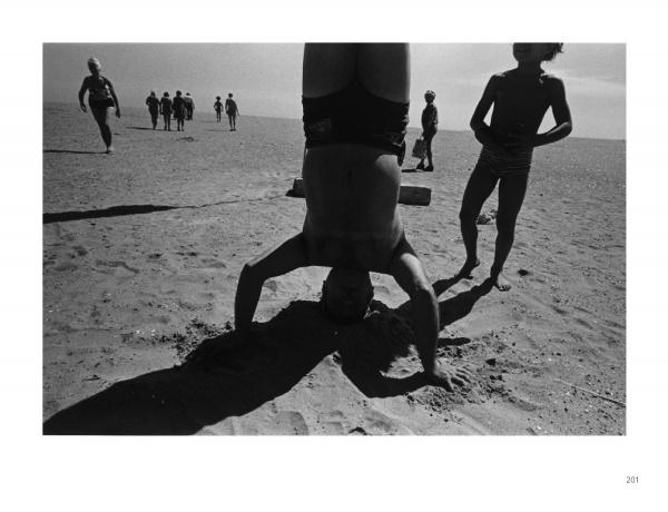 A person balances on their head in the sand at Coney Island, casting a shadow, while children walk nearby under a bright sky—an image from Schiffer Publishing, Ltd.'s "Coney Island," evoking classic black and white photography.