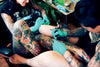 A tattoo artist in green gloves inks a client’s tattooed leg, both adorned with multiple tattoos. This vibrant scene, set in a bright studio, is captured in "AOI Ink Nymphs Los Angeles" by Schiffer Publishing, Ltd.