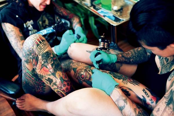 A tattoo artist in green gloves inks a client’s tattooed leg, both adorned with multiple tattoos. This vibrant scene, set in a bright studio, is captured in "AOI Ink Nymphs Los Angeles" by Schiffer Publishing, Ltd.