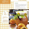 A jar of Schiffer Publishing, Ltd. Squash & Zucchini marmalade with a green ribbon sits on a table next to fresh apricots, a spoon, a cookbook, and an ingredients list in the background.