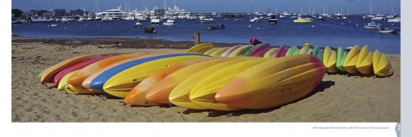 A row of colorful kayaks on a sandy beach with boats in the harbor under a blue sky—an iconic Nantucket scene from "Nantucket Vistas" by Schiffer Publishing, Ltd., perfect for photography lovers or souvenir collectors.