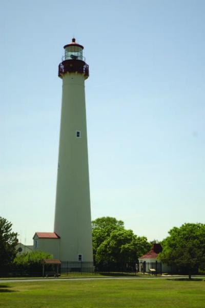 A tall, white lighthouse with a red top stands among trees and small buildings under a blue sky—a highlight featured in Cape May Walking Tours by Schiffer Publishing, Ltd.