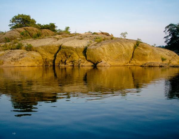 Rocky outcrops with sparse vegetation mirrored in calm blue water under a clear sky—a serene scene reminiscent of the photography in "Faces of the Susquehanna" from Schiffer Publishing, Ltd.