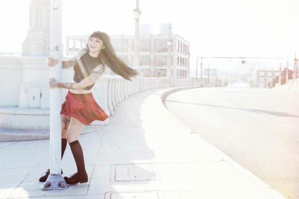 A woman with long dark hair and tattoos, wearing a red skirt and black top, smiles by a street pole in LA—an image capturing the spirit of AOI Ink Nymphs Los Angeles by Schiffer Publishing, Ltd., perfect for tattooed model features.