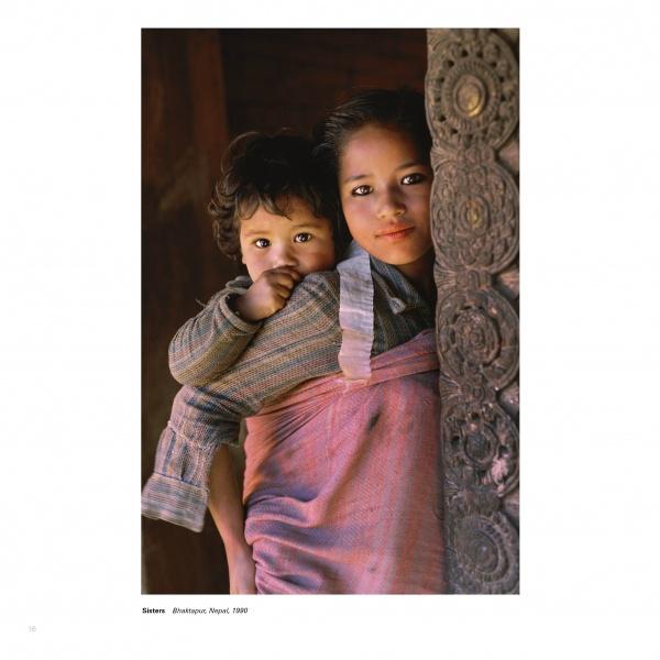 A young girl in “Face to Face” by Schiffer Publishing, Ltd. stands beside a carved post, carrying a child on her back with a pink cloth; both gaze gently at the camera in this striking portrait from Bhaktapur, Nepal, 1980.