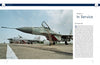 Two MiG-29 air-superiority fighters, featured in "The MiG-29" by Schiffer Publishing, Ltd., are parked on an airfield under a clear sky, with a building and trees visible in the distance. One fighter stands in the foreground and another in the background.