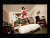 A person wearing a red corset and pink tutu jumps joyfully above a bed in a cozy bedroom, evoking burlesque vibes—capturing the playful spirit of "It's All That Glitters" by Schiffer Publishing, Ltd., among bookshelves and large windows.