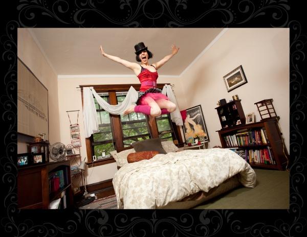 A person wearing a red corset and pink tutu jumps joyfully above a bed in a cozy bedroom, evoking burlesque vibes—capturing the playful spirit of "It's All That Glitters" by Schiffer Publishing, Ltd., among bookshelves and large windows.
