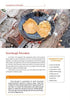 Two pancakes cook in a black grill pan over an outdoor wood fire, as shown in the book "On an Open Fire" by Schiffer Publishing, Ltd. A sourdough pancake recipe and a yellow ingredients box appear below the image.