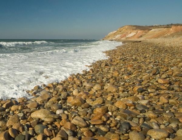 Experience the beauty of Martha’s Vineyard Wide by Schiffer Publishing, Ltd.—smooth stones along a rocky shore lead to ocean waves and a clear sky, with distant cliffs reminiscent of iconic coastal panoramas.