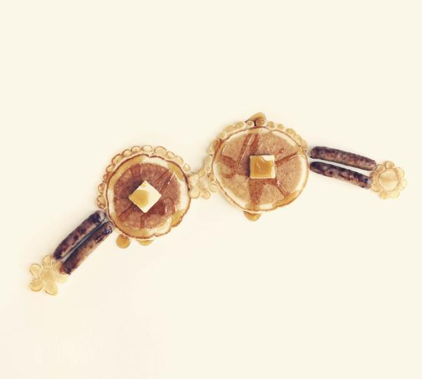 Two pancakes with butter are side by side, with sausage links and syrup dollops forming playful brassieres, evoking a whimsical bra anthology, set against a light background. Product: No Lumps, Thank You by Schiffer Publishing, Ltd.