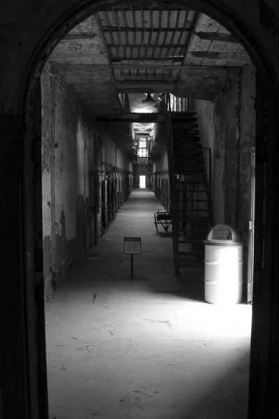 A black and white photo features a long, empty, decaying prison corridor at Eastern State Penitentiary—one of Philadelphia’s haunted places highlighted in Schiffer Publishing, Ltd.’s *Philadelphia Haunts*. Eerie light and barred doors set a chilling scene.