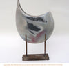 The Pit Firing Ceramics sculpture from Schiffer Publishing, Ltd. features a curved, crescent-shaped form with a marbled white, black, and pink finish on an upright rusted metal base supported by two vertical rods.
