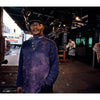 A man in a purple shirt and cap stands in a dim Meatpacking District market, with a butcher and hanging meat behind him. Signs for eggs and meats are visible, referencing the atmosphere featured in "Blood and Beauty" by Schiffer Publishing, Ltd.