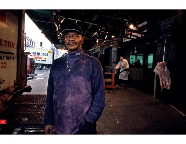 A man in a purple shirt and cap stands in a dim Meatpacking District market, with a butcher and hanging meat behind him. Signs for eggs and meats are visible, referencing the atmosphere featured in "Blood and Beauty" by Schiffer Publishing, Ltd.