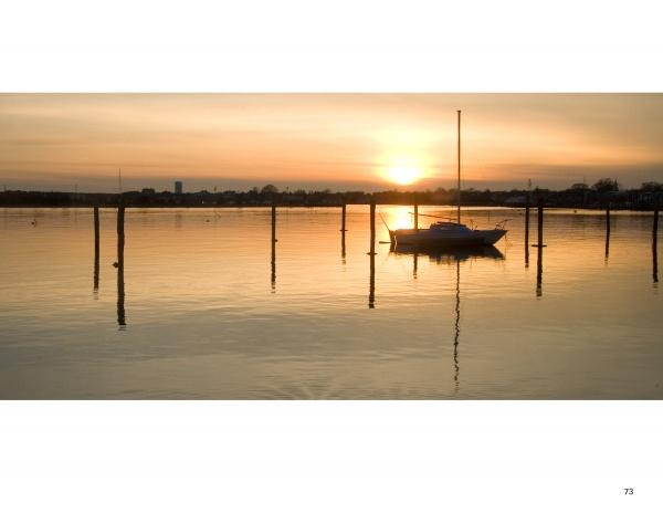 A small sailboat rests on calm water at sunset near Martha’s Vineyard, its reflection shimmering, as captured in "Martha’s Vineyard Wide" by Schiffer Publishing, Ltd., with warm light and wooden posts completing the tranquil scene.