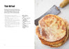 A stack of round, golden-brown gluten-free bread from "The New Bread" by Schiffer Publishing, Ltd. rests on a white surface next to a vintage metal spatula. The rustic, bright image highlights the bread’s texture and the appeal of gluten-free baking.
