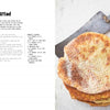A stack of round, golden-brown gluten-free bread from "The New Bread" by Schiffer Publishing, Ltd. rests on a white surface next to a vintage metal spatula. The rustic, bright image highlights the bread’s texture and the appeal of gluten-free baking.