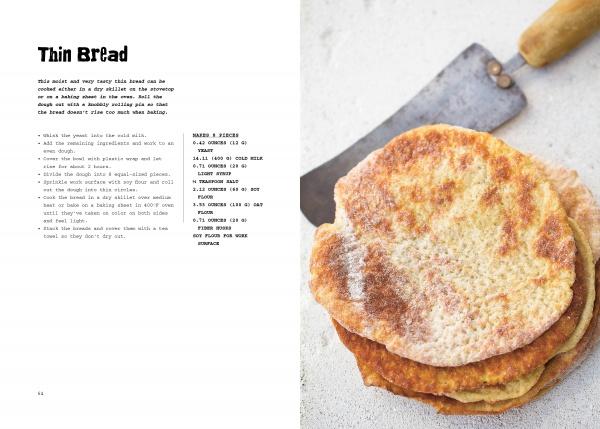 A stack of round, golden-brown gluten-free bread from "The New Bread" by Schiffer Publishing, Ltd. rests on a white surface next to a vintage metal spatula. The rustic, bright image highlights the bread’s texture and the appeal of gluten-free baking.