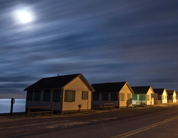 Cape Cod Nights by Schiffer Publishing, Ltd. captures small seaside cabins along a quiet road beneath a cloudy, moonlit sky, evoking the peaceful charm of Cape Cod nights with soft moonlight shimmering on the ocean.