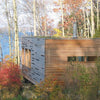 The "Cabin" by Schiffer Publishing, Ltd. is a modern wooden retreat with horizontal slats, nestled near a lake amid autumn trees; its blend of log cabin architecture and contemporary design creates a tranquil, scenic setting.