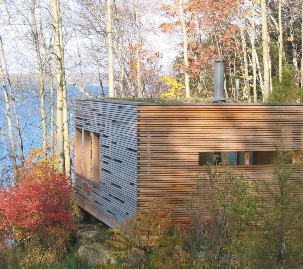The "Cabin" by Schiffer Publishing, Ltd. is a modern wooden retreat with horizontal slats, nestled near a lake amid autumn trees; its blend of log cabin architecture and contemporary design creates a tranquil, scenic setting.