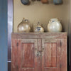 In "Passion for Primitives" by Schiffer Publishing, Ltd., a rustic red wooden cabinet with ceramic jugs and a rack of woven baskets captures the inviting charm of primitive décor in a cozy, vintage setting.