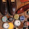 The Leather Care Compendium by Schiffer Publishing, Ltd. is displayed with leather conditioner and brushes around brown leather dress shoes and a single loafer on a wooden surface.