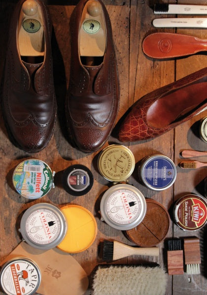 The Leather Care Compendium by Schiffer Publishing, Ltd. is displayed with leather conditioner and brushes around brown leather dress shoes and a single loafer on a wooden surface.