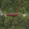 Aerial view of a red covered bridge, like those on the Bridges of Lancaster County Postcards by Schiffer Publishing, Ltd., spanning a narrow brown river amid dense green trees—capturing the timeless charm of classic color postcards.