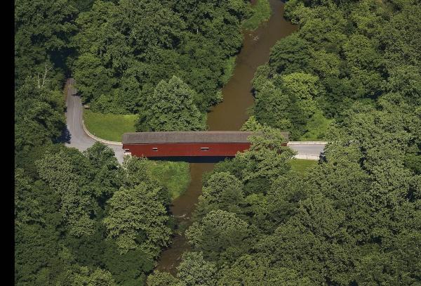 Aerial view of a red covered bridge, like those on the Bridges of Lancaster County Postcards by Schiffer Publishing, Ltd., spanning a narrow brown river amid dense green trees—capturing the timeless charm of classic color postcards.
