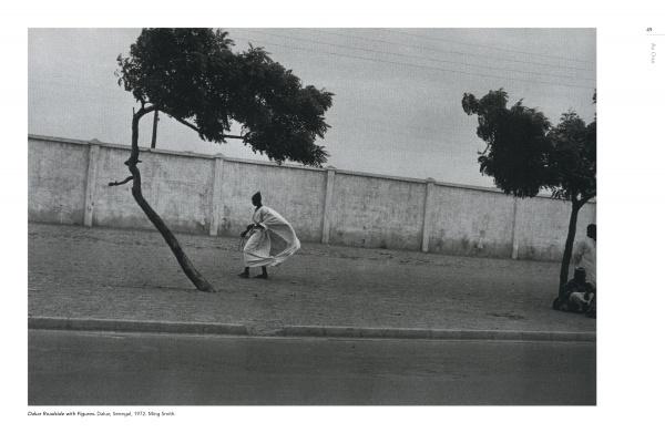 A person in flowing clothing walks beside bent trees and a tall white wall under clouds, echoing scenes from "Timeless" by Schiffer Publishing, Ltd.; another person crouches by a tree at the right edge.