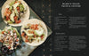 A dark plate holds three vegan tacos with diced tomatoes, lettuce, avocado, black beans, and tempeh. Beside it is Schiffer Publishing, Ltd.’s "Knives and Needles," a cookbook featuring Blake’s home recipes and vegan ceviche.