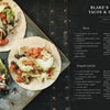 A dark plate holds three vegan tacos with diced tomatoes, lettuce, avocado, black beans, and tempeh. Beside it is Schiffer Publishing, Ltd.’s "Knives and Needles," a cookbook featuring Blake’s home recipes and vegan ceviche.
