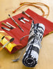 A red fabric brush holder with paintbrushes lies open on a wooden surface next to a rolled-up brush holder featuring a black-and-white cityscape patchwork, crafted using techniques from "Free-Form Patchwork Techniques" by Schiffer Publishing, Ltd.