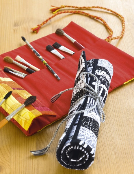 A red fabric brush holder with paintbrushes lies open on a wooden surface next to a rolled-up brush holder featuring a black-and-white cityscape patchwork, crafted using techniques from "Free-Form Patchwork Techniques" by Schiffer Publishing, Ltd.