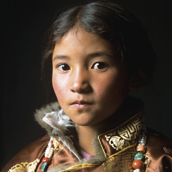 A young girl with brown eyes and wavy hair wears a gold-patterned coat and traditional jewelry, softly gazing at the camera. This striking image is featured in "Ladakh" by Schiffer Publishing, Ltd.