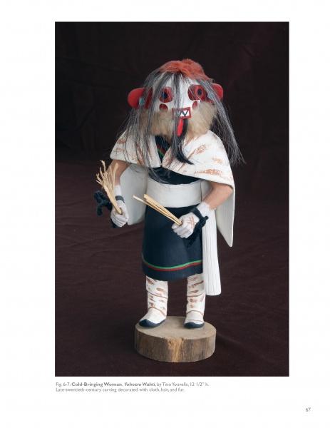 A carved female figure inspired by Hopi Kachinas, wearing a white shawl and dark garment with white trim, fur hair, red and white face paint, holding sticks. Product: Hopi Kachinas by Schiffer Publishing, Ltd.